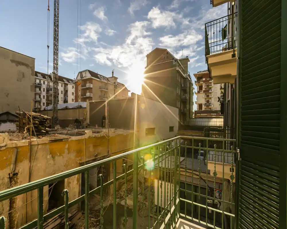 Balcón urbano soleado con vistas a edificios vecinos y una obra; contraventanas verdes y barandilla metálica enmarcan una escena cálida de hora dorada.