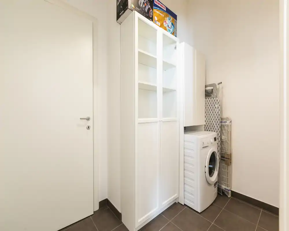 Pequeña zona de servicio/entrada con armario de almacenamiento y lavadora de carga frontal; suelo de baldosa y paredes neutras.