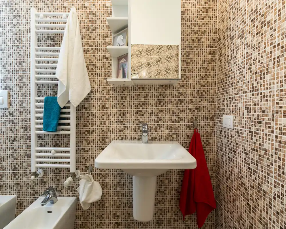 Baño compacto y moderno con paredes de mosaico, lavabo empotrado en la pared y radiador toallero. Los sanitarios limpios y la buena iluminación crean un espacio funcional.