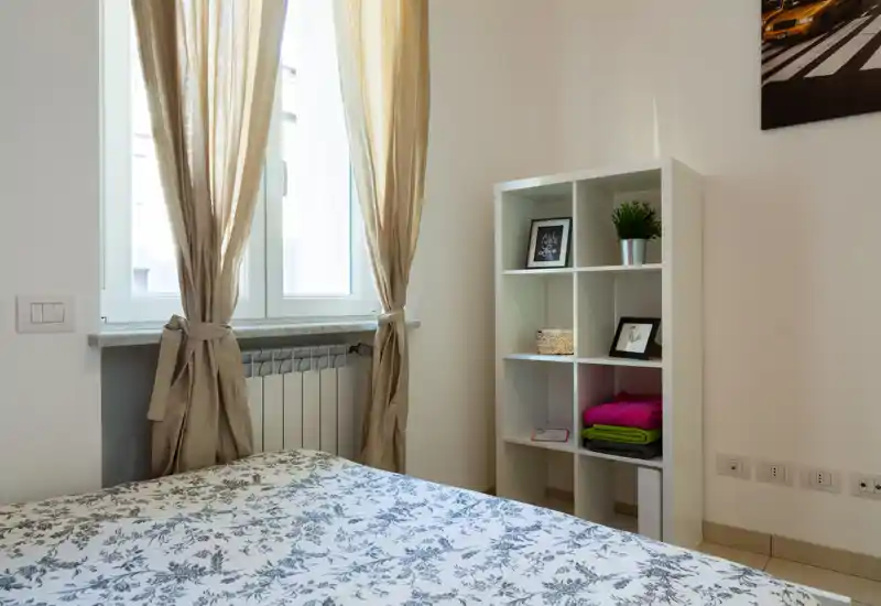 Bright, neatly staged bedroom with a patterned bedspread, light curtains and a compact shelving unit — ideal for cozy short-term stays.
