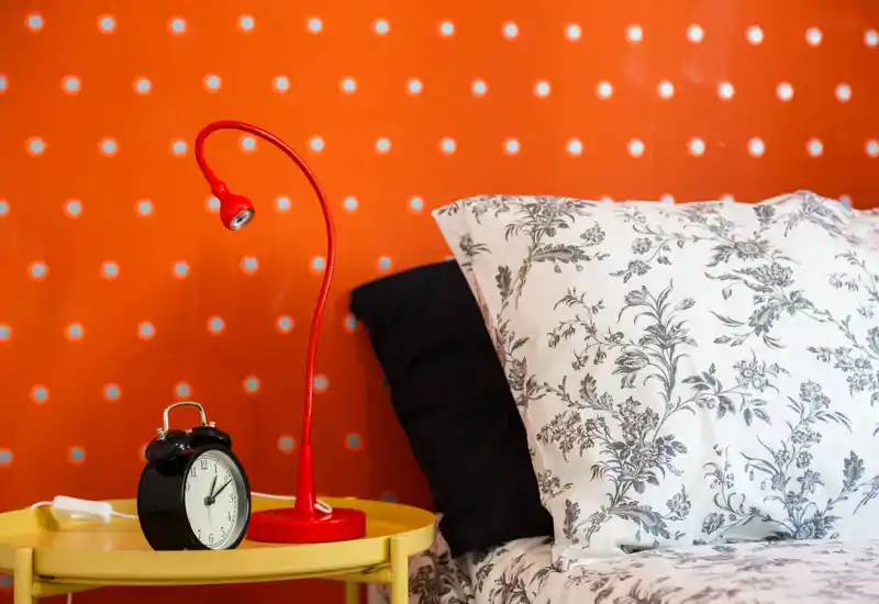 Bright, stylish bedroom detail showing the bed pillows, patterned duvet and a colorful bedside table with lamp and alarm clock — ideal for a cozy night stay.