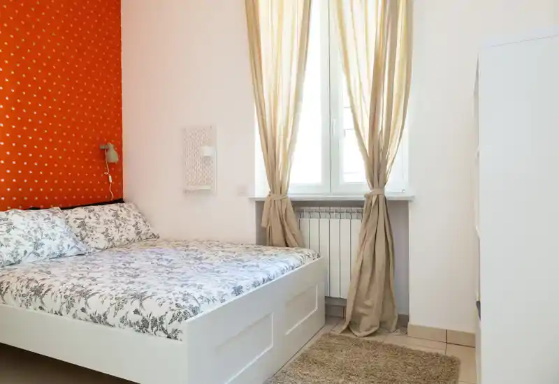 Bright, well-styled bedroom with a white bed frame, patterned bedding and natural light from the window. A warm accent wall and soft textiles create a welcoming atmosphere.