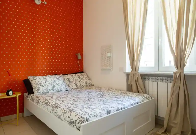 Bright, cozy bedroom with a patterned red accent wall, double bed with floral bedding, and natural light from a large window with beige curtains.
