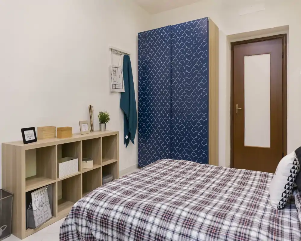 Dormitorio bien presentado con una gran cama a cuadros en primer plano, un armario con diseño y estanterías abiertas para almacenamiento. Luminoso, ordenado y listo para un anuncio de alquiler.