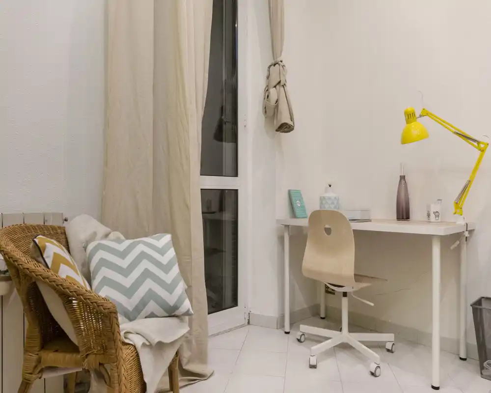 Rincón de estar luminoso y decorado con un sillón de mimbre, cojines con estampado y un pequeño escritorio con silla y lámpara amarilla — un acogedor espacio de trabajo en el salón.