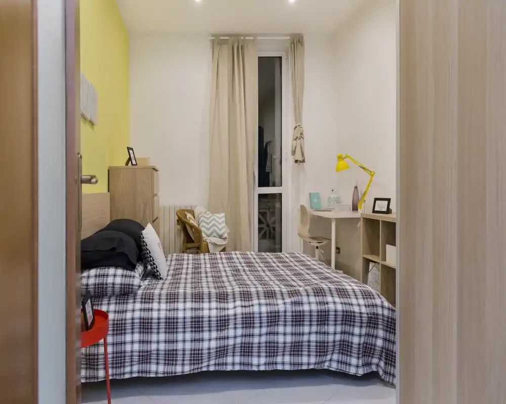 Dormitorio luminoso y bien decorado con cama doble con colcha a cuadros, un escritorio pequeño con lámpara amarilla junto a la ventana y cortinas neutras que crean un ambiente acogedor.