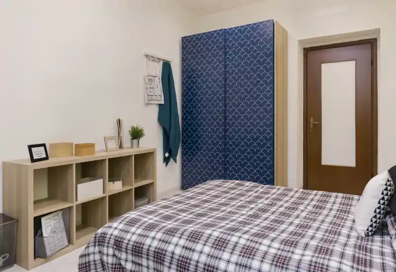 Camera da letto luminosa e ben allestita con armadio decorato e un grande letto con copriletto a quadri. Mensole semplici e tessuti morbidi creano un ambiente accogliente adatto a un annuncio.