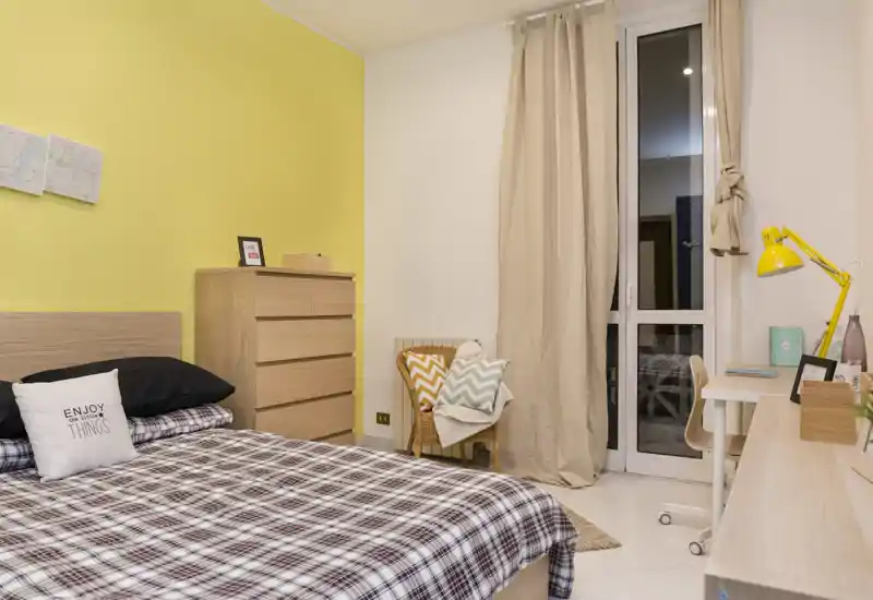 Camera da letto luminosa e ben organizzata con letto matrimoniale, biancheria a quadri e parete d'accento gialla. Presente comò, scrivania e una piccola poltroncina vicino alla finestra con tende.