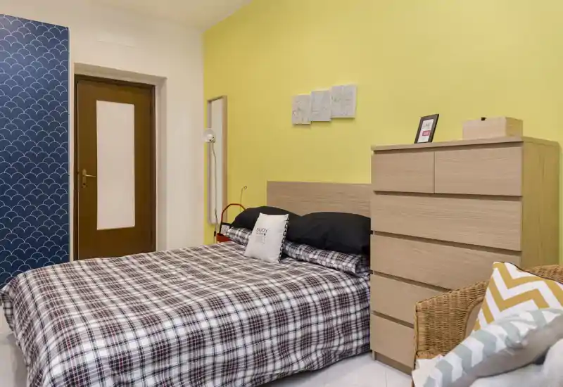 Dormitorio luminoso y bien decorado con cama doble vestida con ropa de cuadros, cómoda de madera y detalles acogedores; ideal para anuncios de alquiler que destacan comodidad y sencillez moderna.