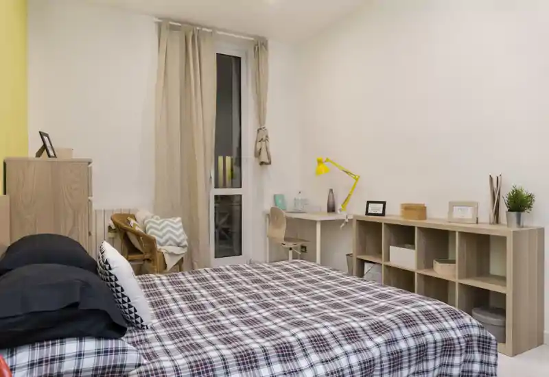 Dormitorio luminoso y bien amueblado con una cama doble grande con edredón a cuadros, un escritorio con una lámpara amarilla y estanterías abiertas para almacenamiento — ideal para una persona o pareja que busca un espacio acogedor y funcional.