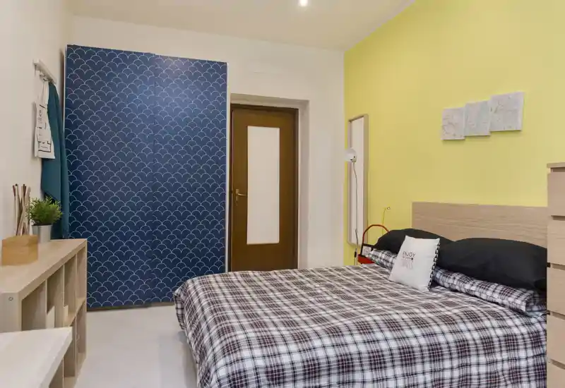 Dormitorio luminoso y ordenado con panel de armario azul estampado, pared amarilla y cama doble con ropa de cama a cuadros. La habitación está bien ambientada con almacenaje, cojines y luz suave, perfecta para anuncios.