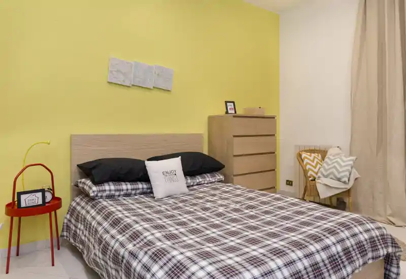 Dormitorio luminoso y decorado con cama doble con ropa de cuadros, cojines decorativos, cabecero de madera y un alto aparador — acogedor y bien iluminado para estancias cortas.