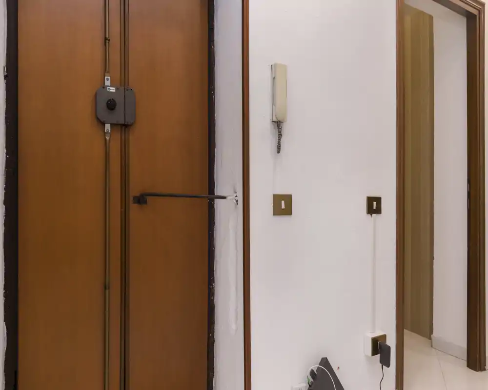 Pequeño recibidor con puerta de madera, portero eléctrico en la pared e interruptor — útil para describir el acceso y las características de seguridad.