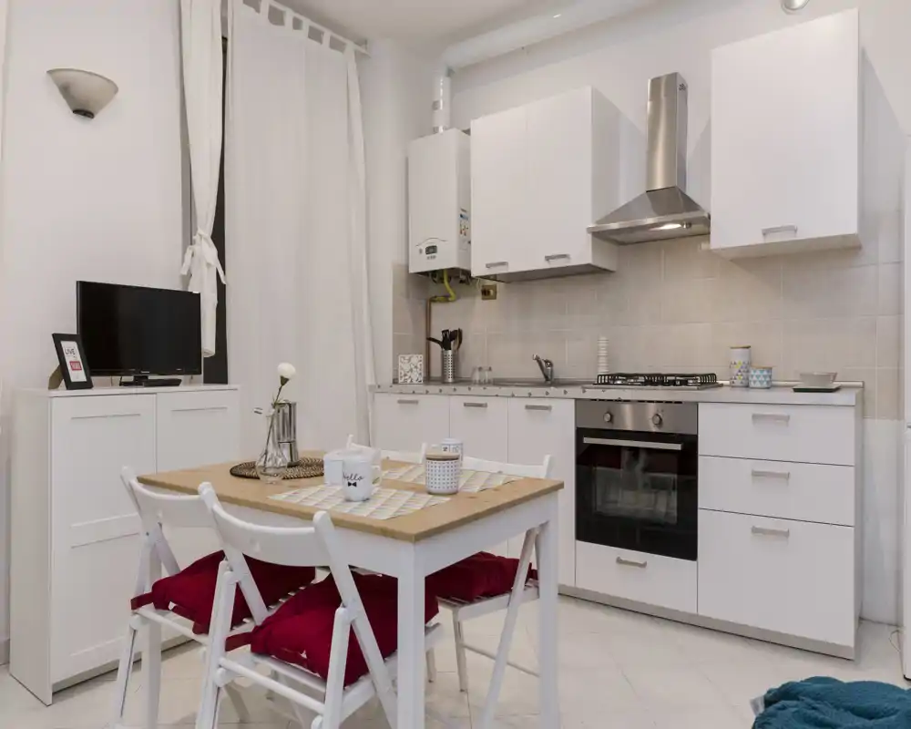 Cocina pequeña y luminosa con armarios blancos, horno empotrado y una mesa de comedor con cojines rojos — ideal para un apartamento compacto.