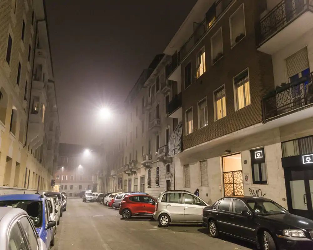 Vista de una calle nocturna con coches aparcados y edificios residenciales bajo farolas, con niebla ligera.