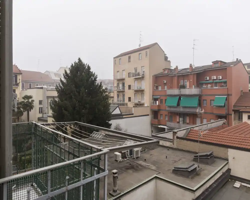 Vistas desde un balcón urbano con tejados y edificios vecinos y una barandilla; luz suave y tonos apagados que aportan una atmósfera tranquila de ciudad.