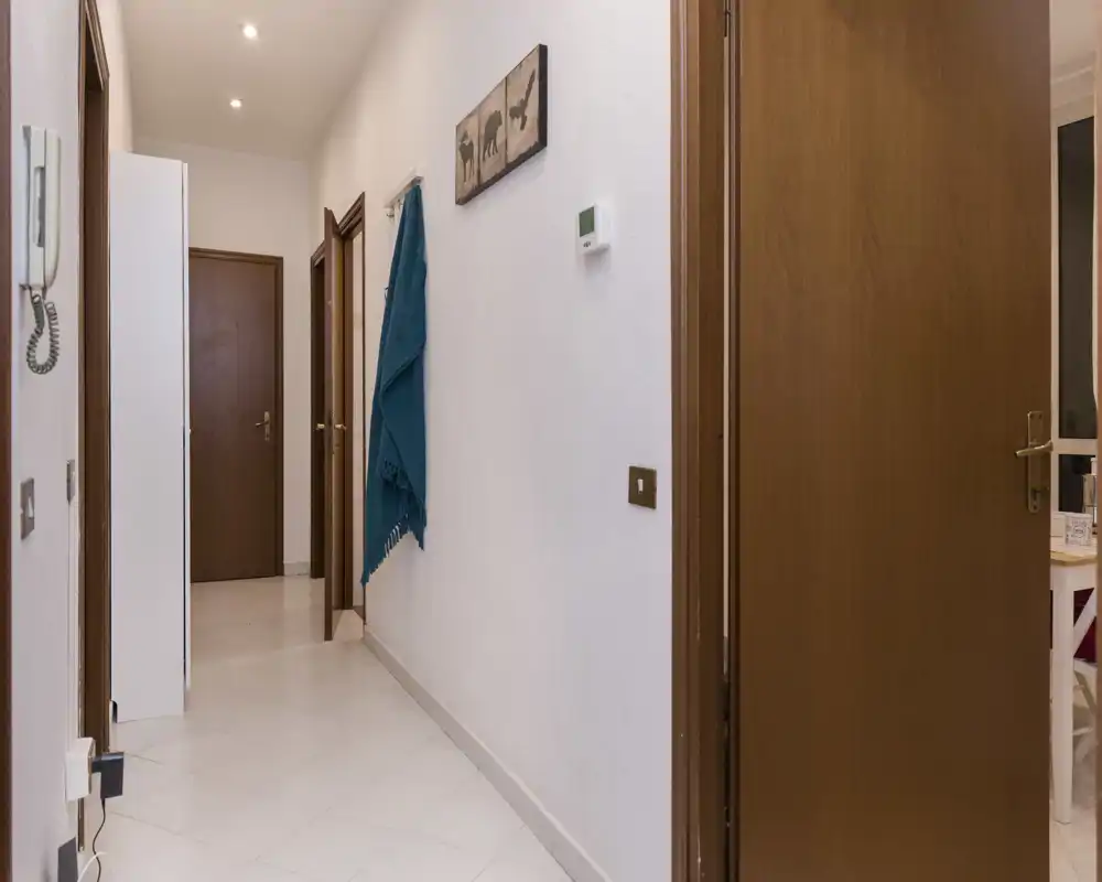 Entrada/pasillo limpio y bien iluminado con paredes blancas, suelo de baldosa y puertas de madera que conducen a otras habitaciones. Un colgador turquesa y un pequeño cuadro aportan personalidad.