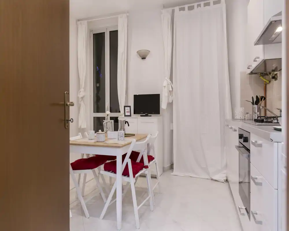 Cocina compacta y luminosa con mobiliario blanco moderno, vitrocerámica y horno a la derecha, y una mesa con sillas con cojines rojos en el centro — espacio ordenado y bien iluminado, ideal para grupos pequeños.