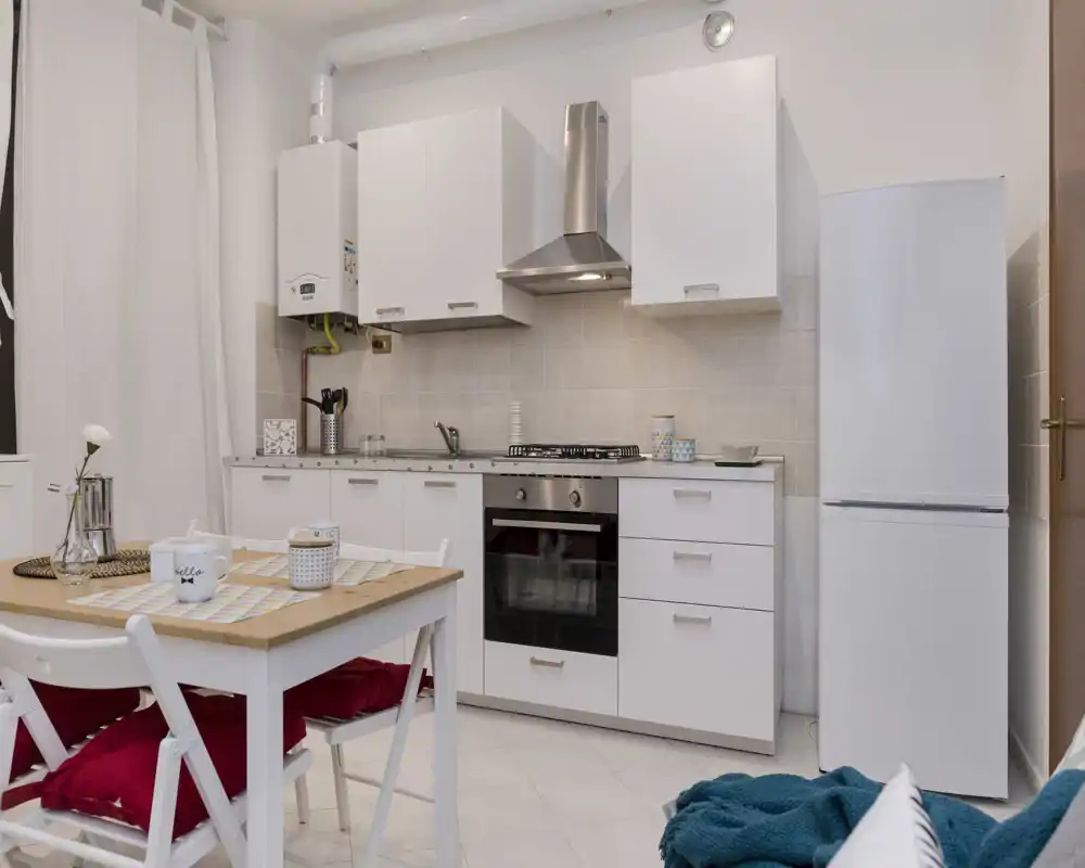 Cocina moderna y compacta con armarios blancos, horno empotrado y frigorífico independiente; pequeña mesa de madera con cojines rojos que crea un acogedor rincón para desayunar.