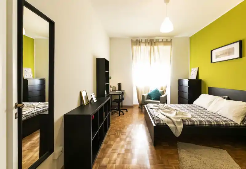 Bright, modern bedroom with a queen bed, soft textiles and a work corner by the window. Warm green accent wall and wooden parquet make the room inviting and airy.