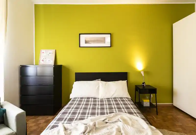 Bright, modern bedroom with a double bed centered against a vivid green accent wall, bedside table with lamp, chest of drawers and cozy textiles.
