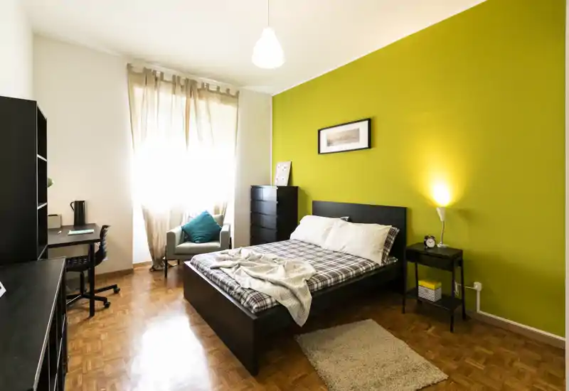 Bright, stylish bedroom with a queen bed centered against a vibrant green accent wall, bedside table with lamp, a chest of drawers and a small work corner by the window.