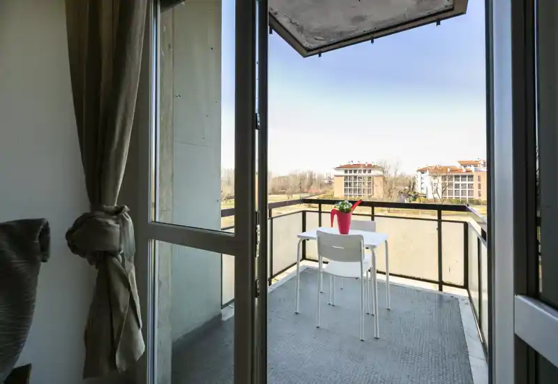 Piccolo balcone soleggiato con un tavolino e una sedia, con vista sugli edifici e gli alberi vicini — perfetto per il caffè mattutino.