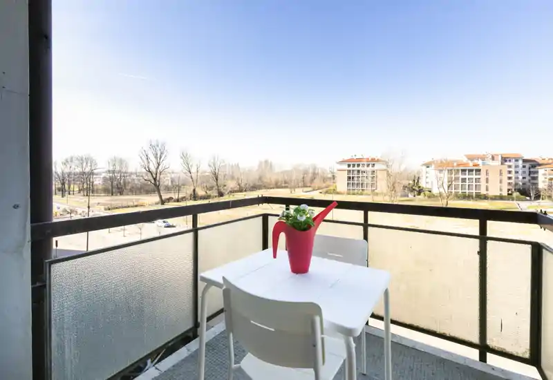 Balcone soleggiato con un piccolo tavolo bianco e due sedie, decorato da un annaffiatoio rosso usato come vaso e con ampia vista su alberi e palazzi vicini.