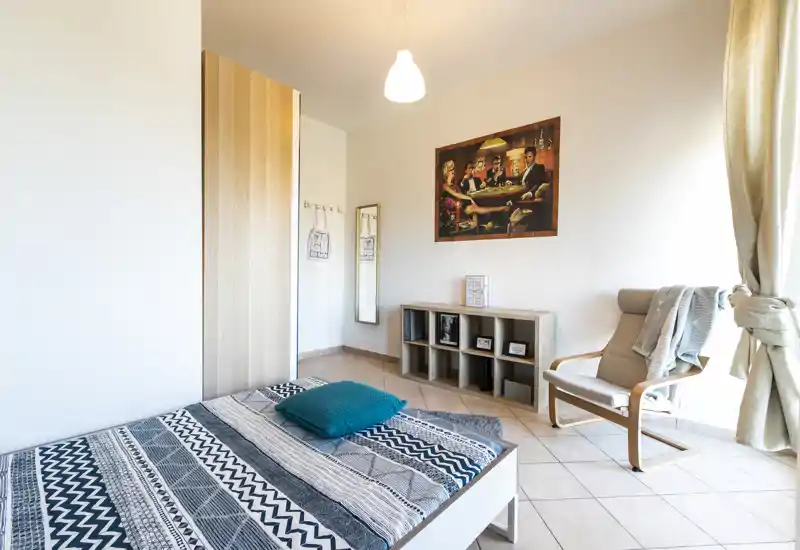 Dormitorio luminoso y ordenado con colcha de estampado, cojín turquesa y un sillón acogedor junto a la ventana — decoración moderna y minimalista, ideal para alquileres.