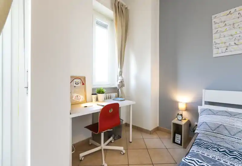 Rincón de dormitorio luminoso y ordenado con cama individual, ropa de cama estampada y pequeño escritorio junto a la ventana — ideal para estudiante o huésped individual.