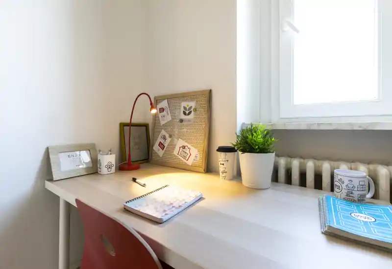 Zona scrivania luminosa e ordinata con lampada da tavolo calda, quaderno, pianta in vaso e luce dalla finestra — perfetta per uno studio o angolo lavoro.