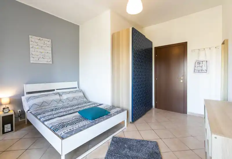 Dormitorio moderno y luminoso con cama doble, ropa de cama estampada, lámpara de mesita y armario sencillo — ordenado y listo para huéspedes.