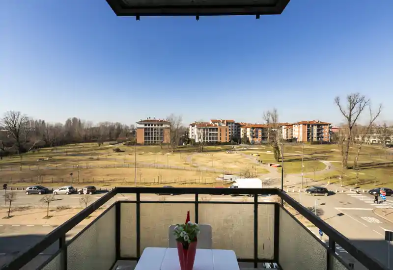 Balcone privato soleggiato con tavolo e composizione floreale, vista su un parco e palazzi residenziali — ideale per la colazione e momenti all'aperto.