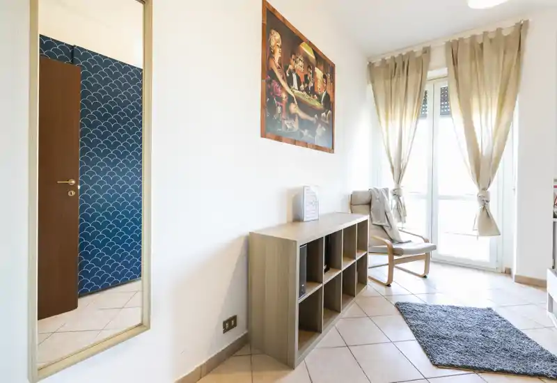 Zona giorno luminosa e minimale con poltrona, libreria bassa e ampia finestra con tende che si affaccia su un balcone — perfetta per rilassarsi con luce naturale.