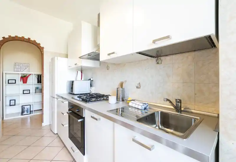 Cucina moderna e luminosa con lavello in acciaio, piano cottura a gas e forno incassato; pensili bianchi e rivestimento a piastrelle per un ambiente pulito e funzionale.