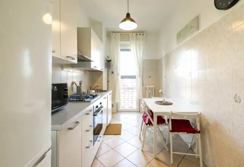Cucina moderna e luminosa a corridoio con pensili bianchi, piano cottura a gas e un piccolo tavolo da pranzo vicino alla porta vetrata che illumina l'ambiente.