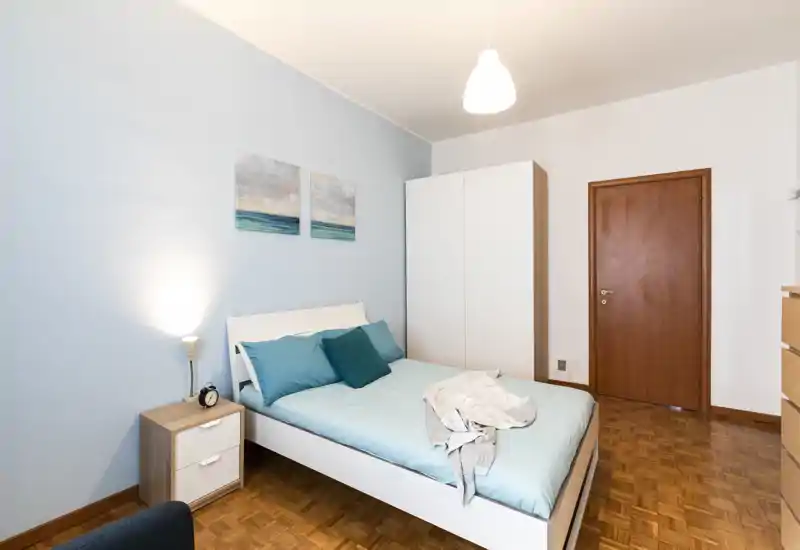 Dormitorio moderno y luminoso con cama doble vestida con lencería turquesa, cojines a juego y armario blanco; iluminación suave y cuadros costeros crean un ambiente tranquilo y acogedor.