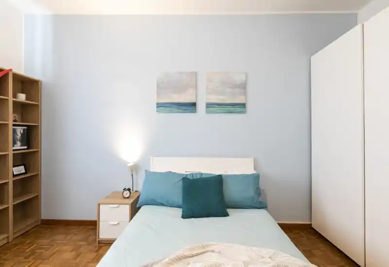 Dormitorio luminoso y ordenado con cama doble centrada en una pared azul clara, cojines turquesa, mesita con lámpara y armario — un espacio para dormir tranquilo y acogedor.