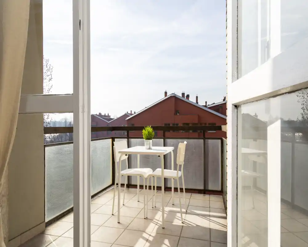 Sunny furnished balcony with a small table and two chairs, tiled floor and a view over neighboring red-roofed buildings. Ideal for morning coffee in bright natural light.