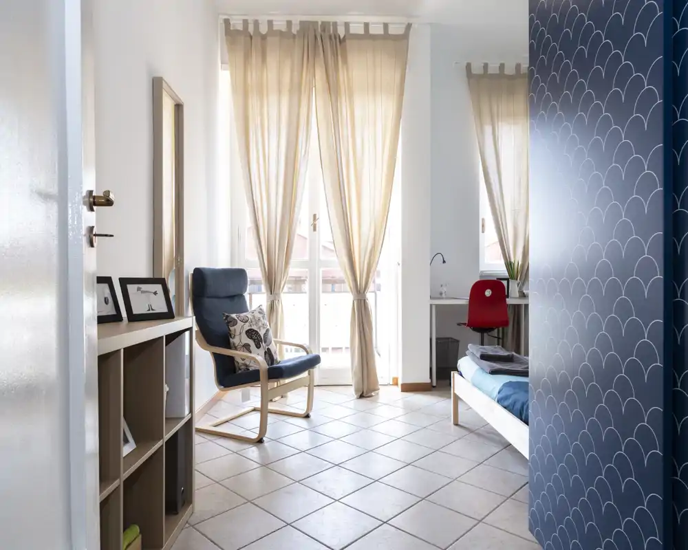 Bright, neatly arranged bedroom with a single bed, reading chair and desk by large sunlit windows — airy, modern and well staged for rental photos.