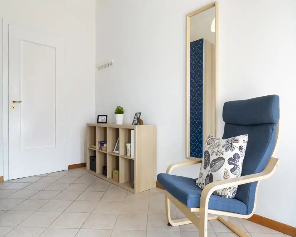 Minimal, bright living area with a comfortable blue armchair, decorative cushion, wall mirror and low wooden shelving—simple, uncluttered and welcoming.