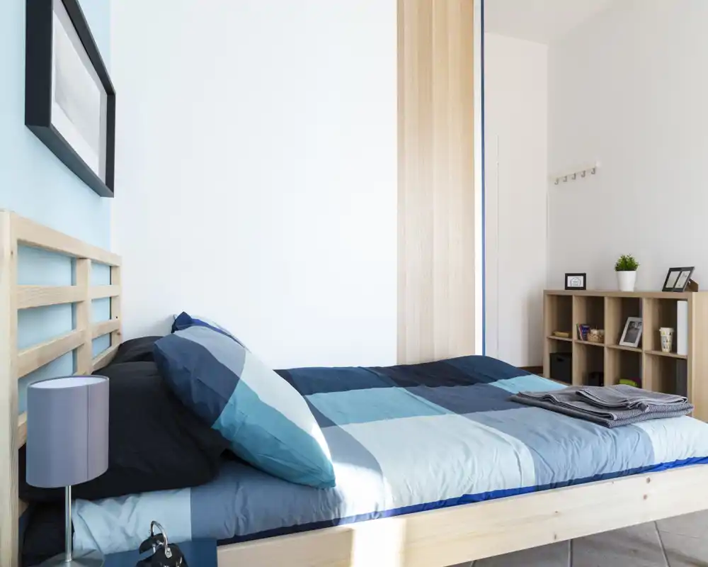 Bright, modern bedroom with a wooden bed frame and blue bedding, neat shelving and natural light creating a fresh, welcoming atmosphere.