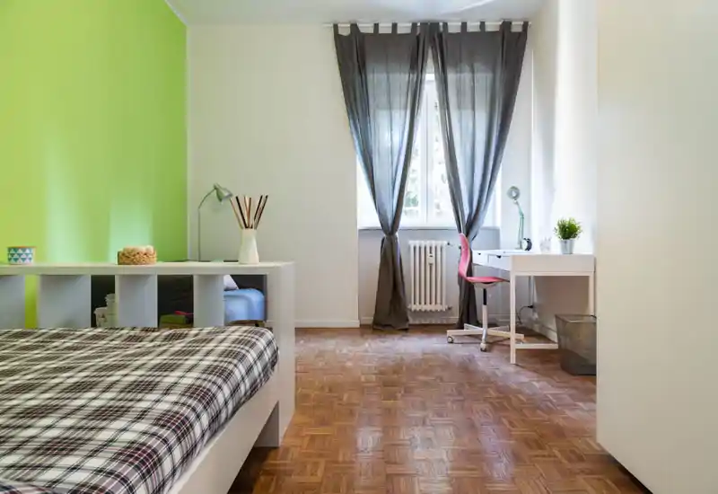 Bright, tidy bedroom with a plaid bed in the foreground, a desk by the window and a green accent wall that adds character.