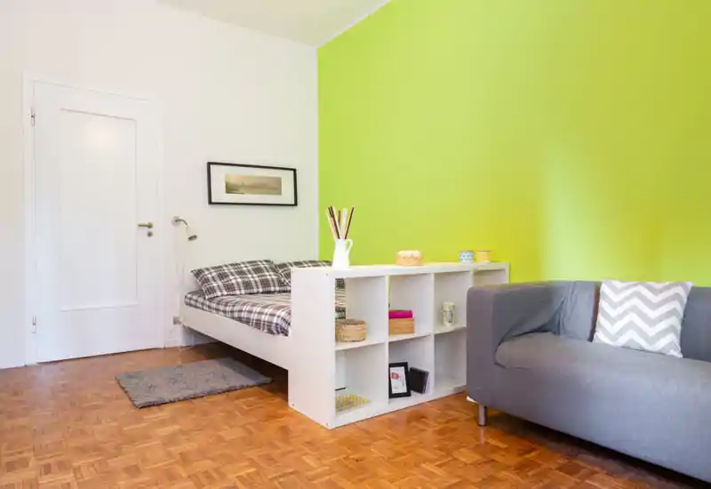 Compact, bright bedroom with a patterned double bed and a small shelving unit acting as a room divider; contemporary grey sofa and lively green accent wall add personality.