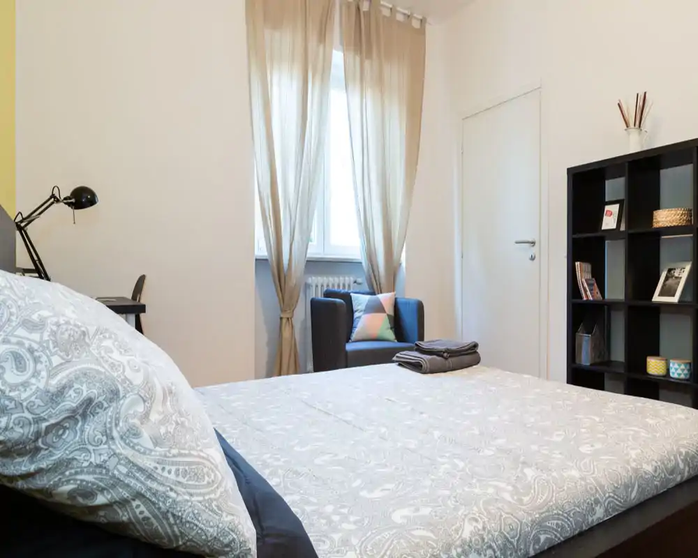 Bright, well-styled bedroom with a large bed in the foreground, an armchair by the window and a shelving unit for storage—ideal for a cozy stay.