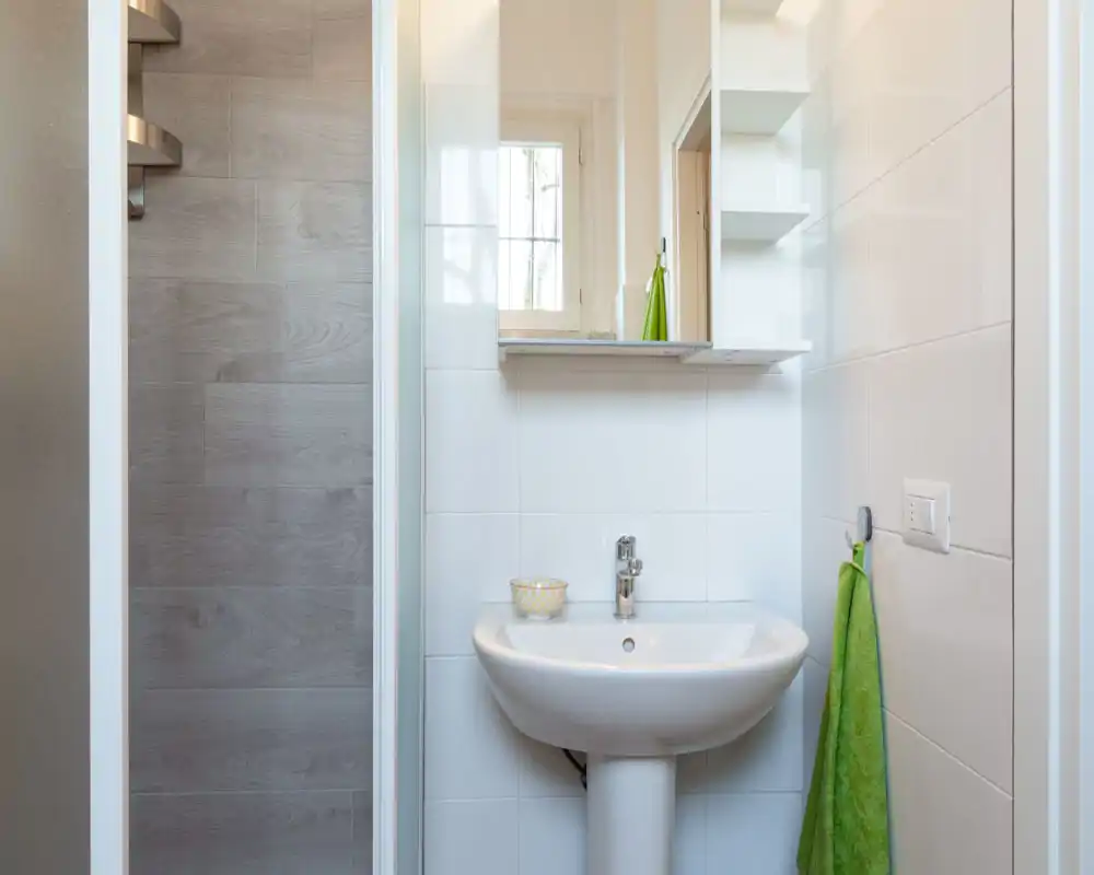 Bright, clean bathroom with a pedestal sink, wall mirror and green towel; compact modern shower visible to the left.