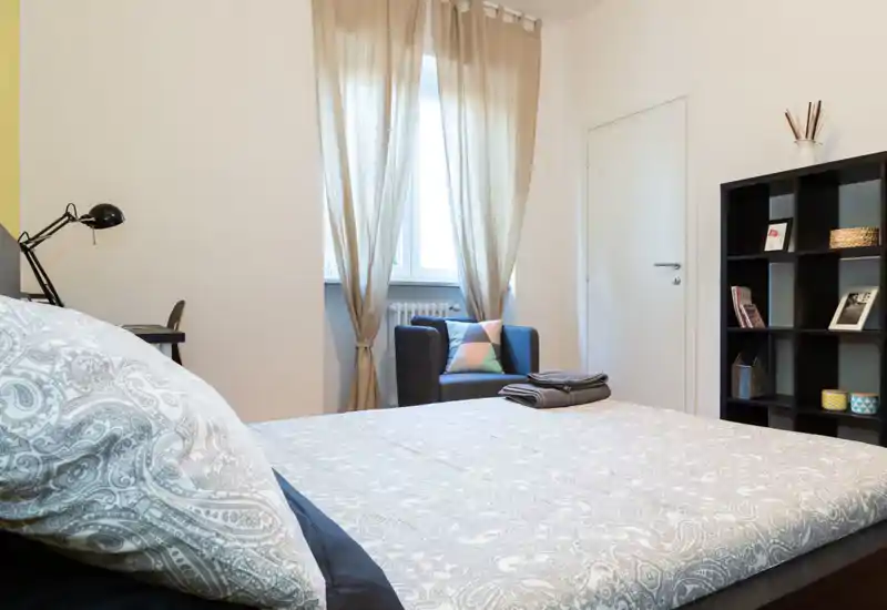 Bright, well-styled bedroom with a large bed in the foreground, an armchair by the window and a shelving unit for storage—ideal for a cozy stay.