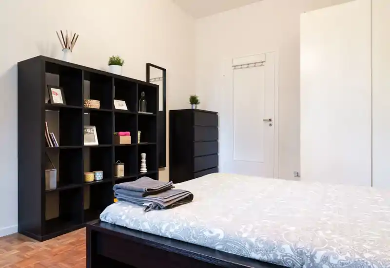 Bright, modern bedroom with a large bed in the foreground, patterned bedding and neatly folded towels; black shelving and dresser provide stylish storage and decor.