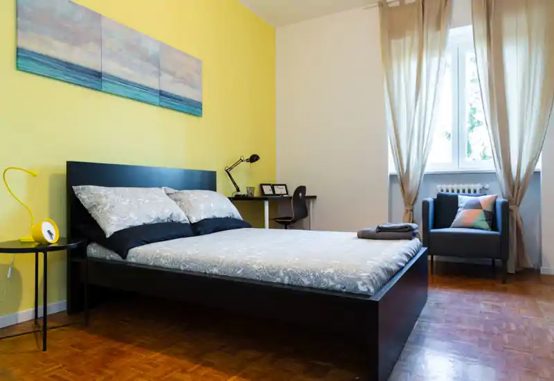 Bright and modern bedroom with a double bed in the center, patterned bedding, a bedside table with lamp, a desk in the corner and a cozy armchair by a large window letting in natural light.