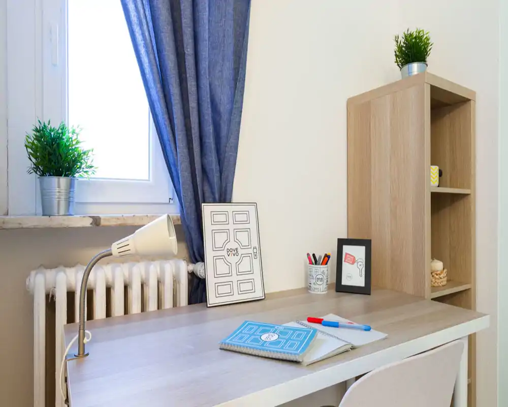 Angolo scrivania luminoso e ordinato vicino alla finestra con lampada, piccolo scaffale e piante decorative — un accogliente spazio studio ideale per smart working o studio.
